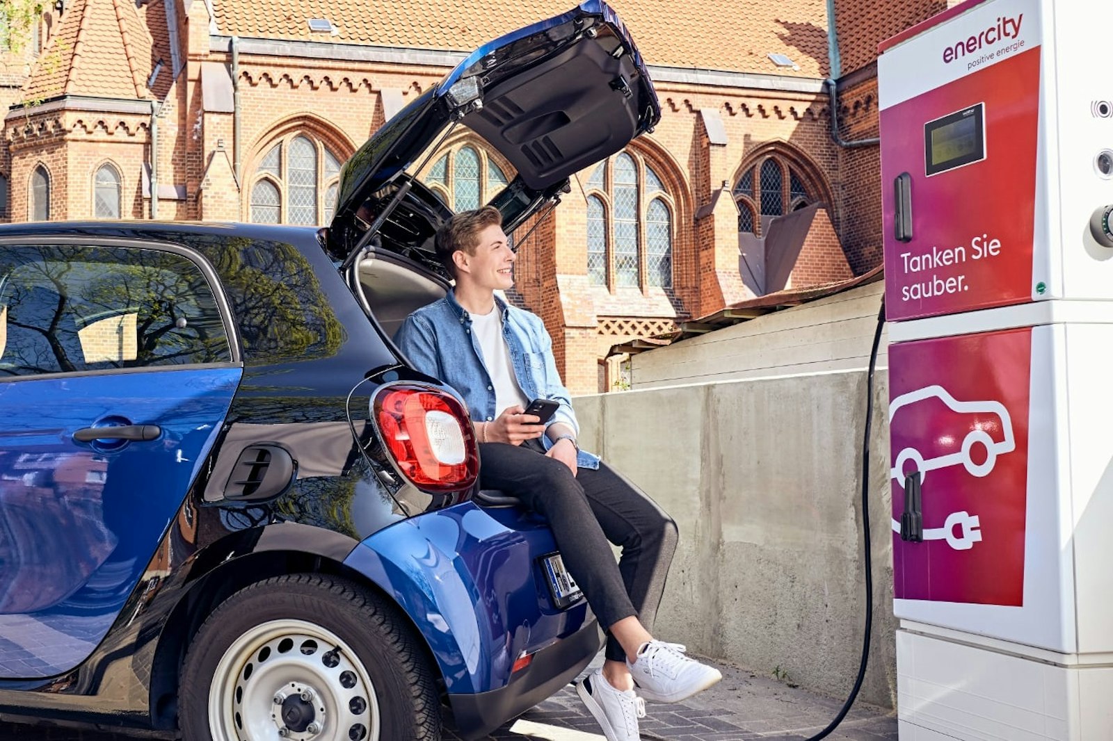 Ladesaeulen-Sharing_TEASER Mann sitzt im Kofferraum seines E-Autos, das an einer Ladebox vor einer Kirche lädt