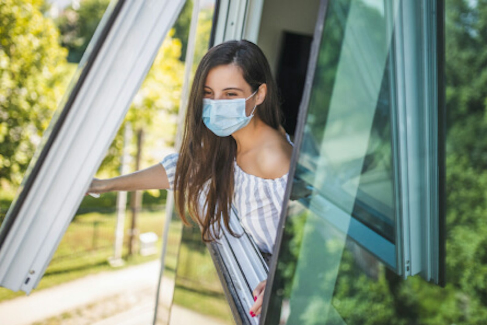 hygienekonzept-an-schulen-lueften.jpg Hygienekonzept. Dieses Bild zeigt eine junge Frau, die eine medizinische Maske auf hat und gerade ein Fenser öffnet.